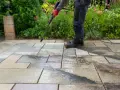 Persona con guantes rojos usa hidrolimpiadora para limpiar suelo de terraza exterior con baldosas de piedra.