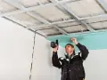 Un obrero instala un **pladur techo** con una taladradora. La estructura metálica ya está fijada al techo original.