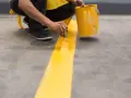 Persona pintando una línea amarilla sobre suelo de cemento sin pulir, con una lata de pintura al lado.
