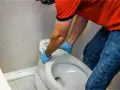 Hombre con guantes azules desmonta asiento de inodoro. Aprende como quitar un bidet para renovar tu baño.