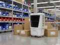 Un climatizador evaporativo blanco con ruedas, rodeado de cajas de cartón y estanterías con contenedores azules y rojos en un almacén.