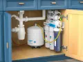 Sistema de ósmosis inversa instalado bajo el fregadero. Un tanque blanco y varios filtros de agua listos para instalar osmosis en casa.