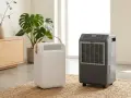 Dos tipos de deshumidificadores, uno blanco y portátil con asa de madera, otro gris con ruedas, en una sala de estar moderna.