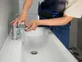 Man con mono azul trabajando para cambiar grifo lavabo. Manos ajustando la base del grifo cromado sobre el lavabo blanco.