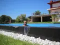 Niña sonriente en una piscina desbordante, con agua cayendo sobre piedras blancas y césped verde. Casa moderna al fondo.