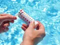 Manos sostienen una tira reactiva para medir el pH de la piscina. El agua azul y brillante de la piscina se ve al fondo.