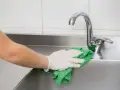 Mano con guante limpiando fregadero de acero inoxidable con paño verde.