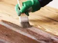 Mano con guante verde aplicando barniz a madera. El proceso de barnizar madera protege y embellece la superficie.