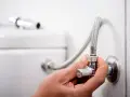 Mano ajustando uno de los tipos de llaves de paso de agua, conectada a una manguera flexible y al tanque de un inodoro.