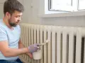 Hombre con barba pintando radiadores blancos con brocha y pintura.
