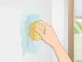 Mano limpiando una mancha en pared blanca con esponja y agua. Aprende como limpiar paredes blancas fácilmente.