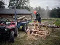 Hombre corta leña con motosierra, pensando cuál es la mejor leña para chimenea. Remolque con troncos y ATV al lado.