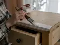 Persona con camisa a cuadros pinta madera de un mueble con brocha blanca.