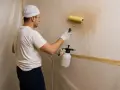 Hombre con rodillo y pulverizador mojando la pared para quitar gotele con agua.