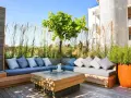 Elegante decoracion terrazas aticos con sofás de madera, cojines grises y un centro de mesa con fuego. Hierbas altas y un árbol crean un oasis urbano.