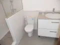 Baño moderno con ducha separada por muro de azulejos blancos y cristal. Inodoro y lavabo sobre mueble blanco.