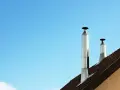 Dos tubos de chimenea metálicos se alzan sobre un tejado marrón contra un cielo azul claro. Es un día perfecto para aprovechar el calor del tubo de chimenea.