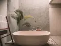 Un baño moderno con una bañera ovalada, toallas blancas en una escalera de madera y una planta de hojas verdes. La pared tiene textura y el suelo está cubierto de guijarros. ¡Listo para pintar la bañera y renovar el espacio!