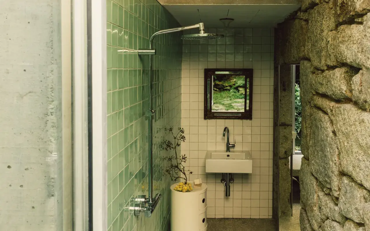 Baños pequeños con ducha, lavabo y espejo con vista a un jardín rocoso. Paredes de azulejos verdes y piedra natural.