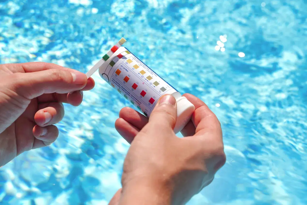 Manos sostienen una tira reactiva para medir el pH de la piscina. El agua azul y brillante de la piscina se ve al fondo.