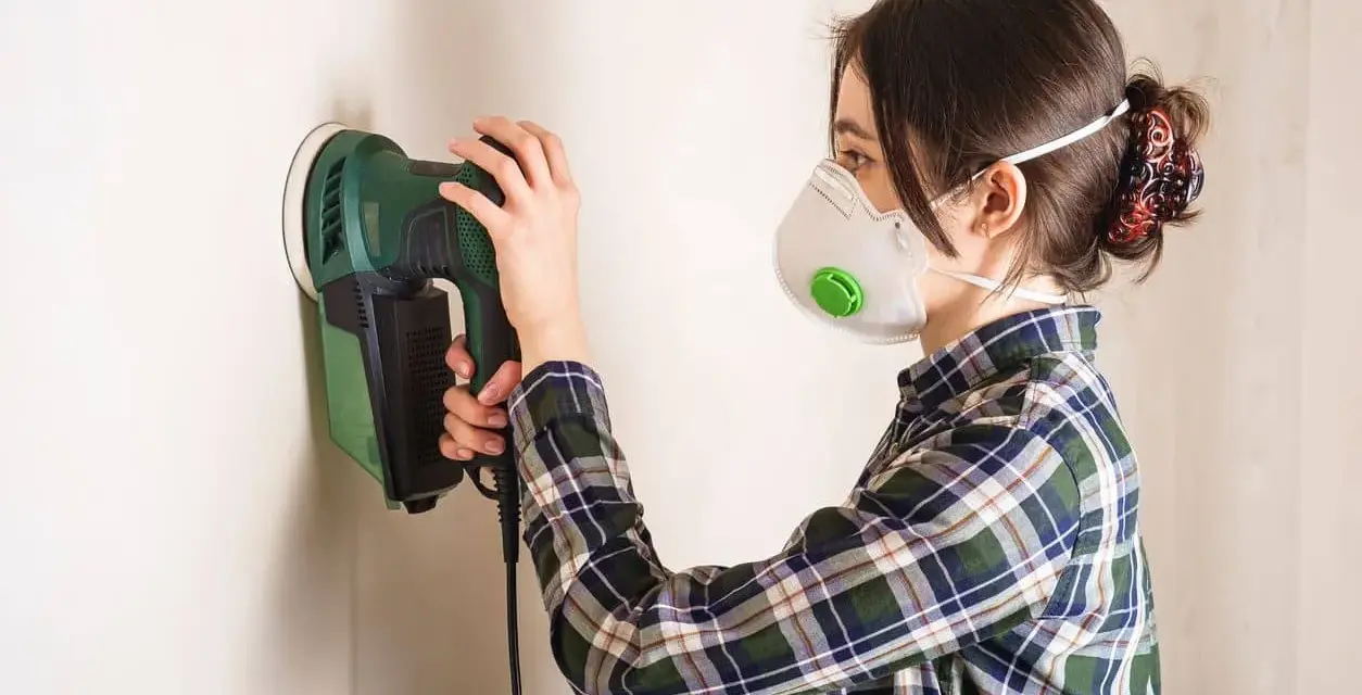 Mujer con mascarilla lijando paredes con una lijadora eléctrica.