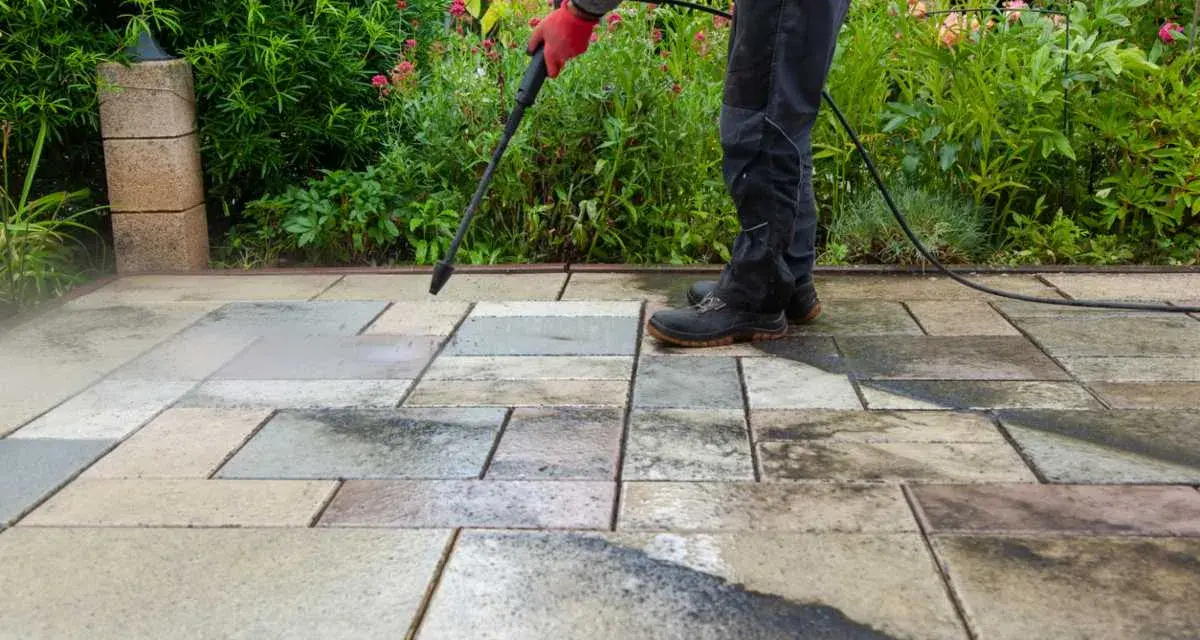 Persona con guantes rojos usa hidrolimpiadora para limpiar suelo de terraza exterior con baldosas de piedra.