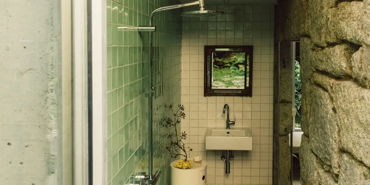 Baños pequeños con ducha, lavabo y espejo con vista a un jardín rocoso. Paredes de azulejos verdes y piedra natural.