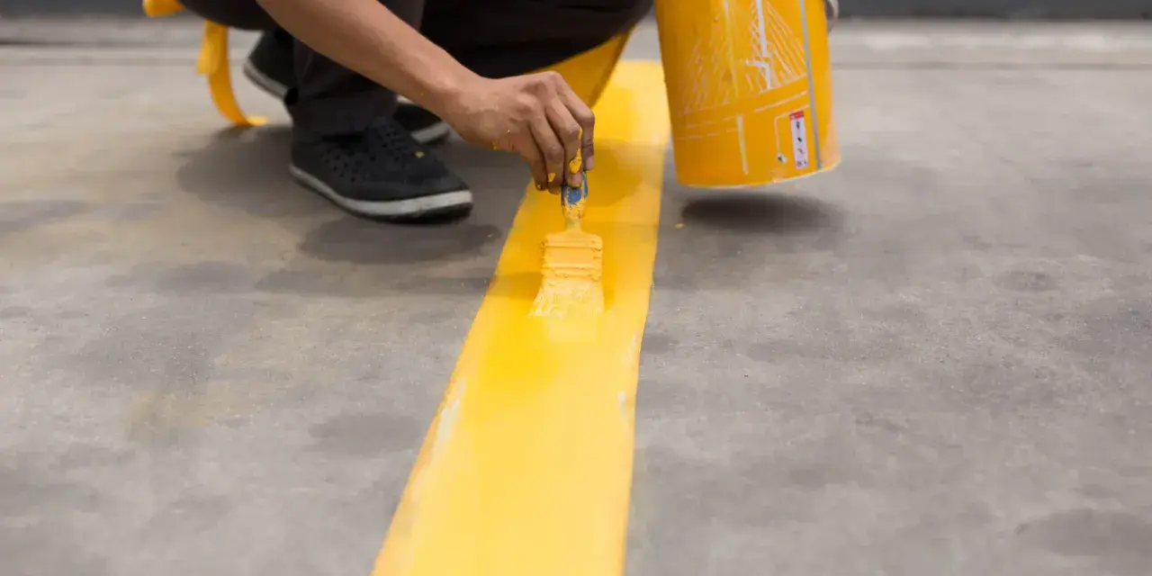Persona pintando una línea amarilla sobre suelo de cemento sin pulir, con una lata de pintura al lado.