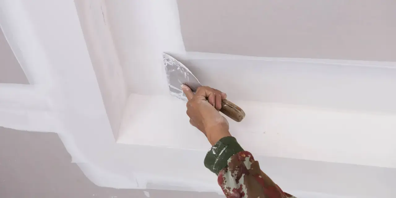 Mano con espátula aplica masilla para reparar techo escayola, dejando un acabado liso y perfecto.