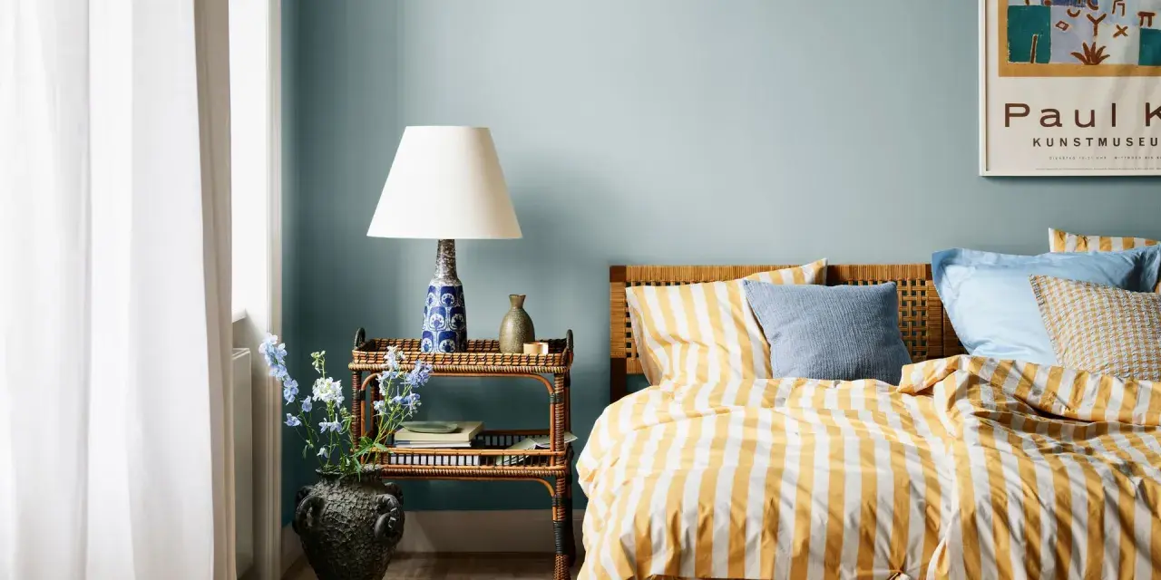 Habitación con paredes azul claro, cama con ropa de cama a rayas amarillas y blancas, y mesita de noche de ratán. Ideas de colores para habitaciones.