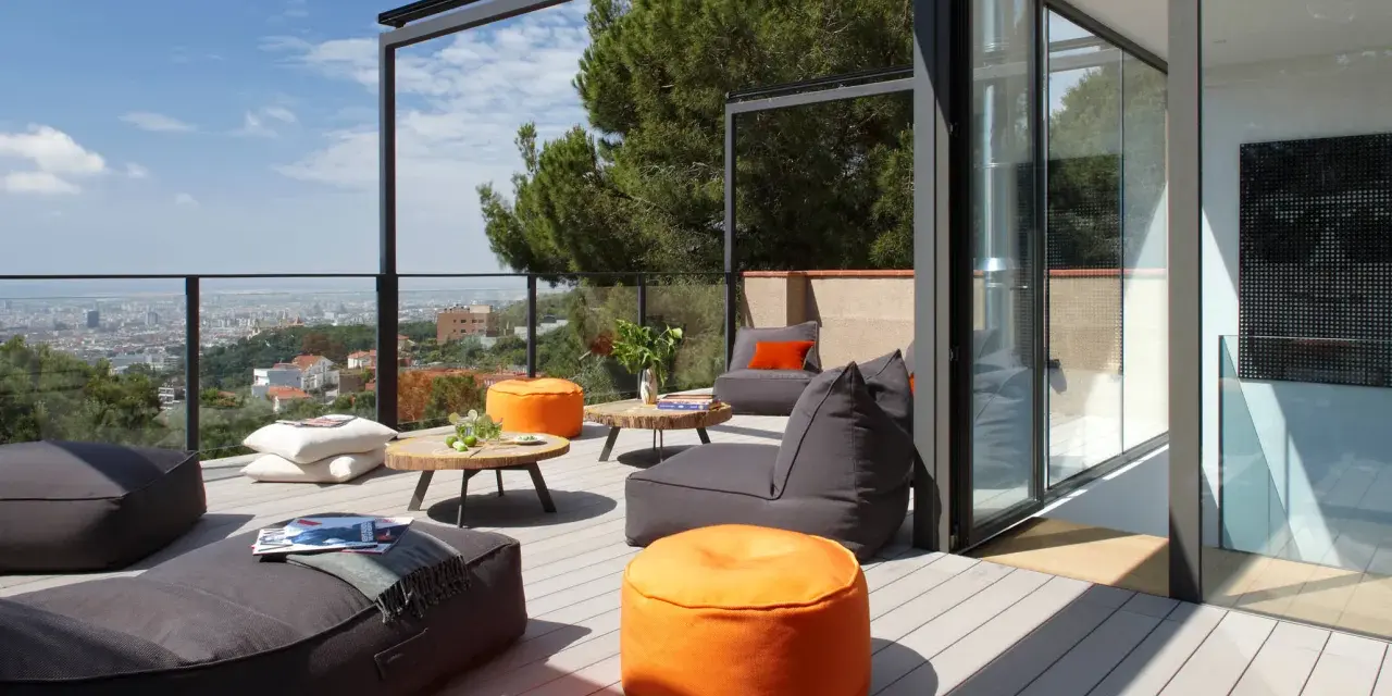 Cómoda terraza ático con sillones y pufs naranjas, vistas a la ciudad y un gran pino.