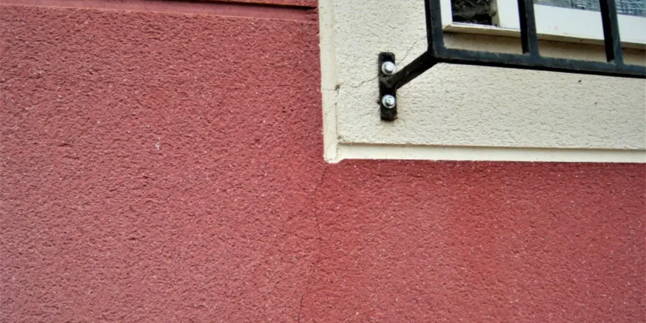 Grietas profundas en pared de color rojo, cerca de una ventana con rejas. Aprende como arreglar grietas profundas en paredes.