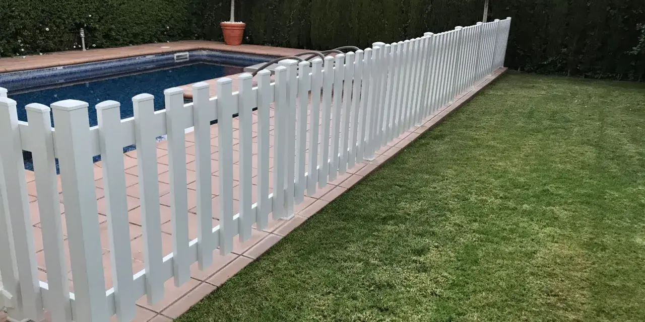 Una verja de jardín blanca protege una piscina y un césped verde.