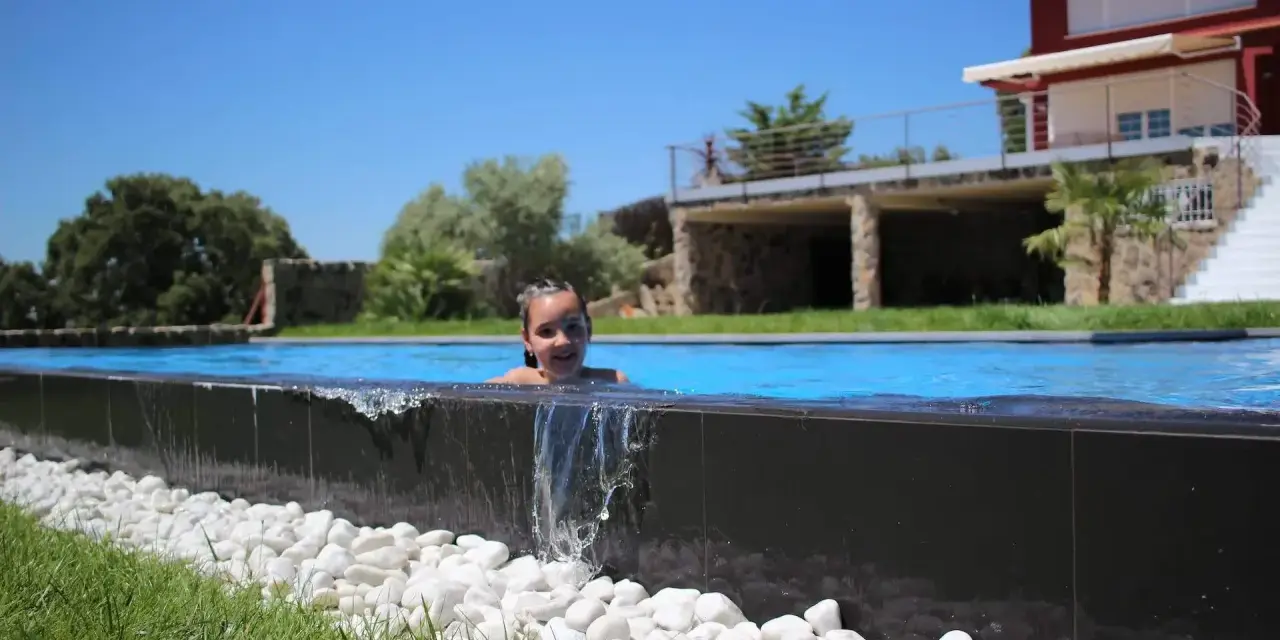 Niña sonriente en una piscina desbordante, con agua cayendo sobre piedras blancas y césped verde. Casa moderna al fondo.