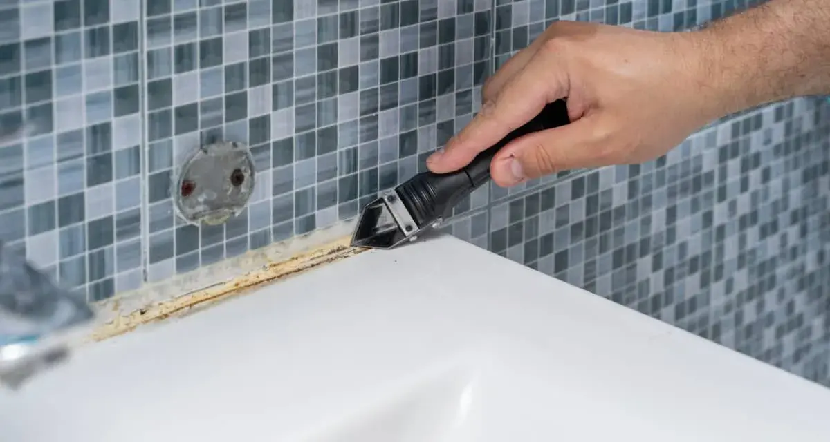 Mano usando una espátula para quitar silicona vieja y sucia de la bañera.
