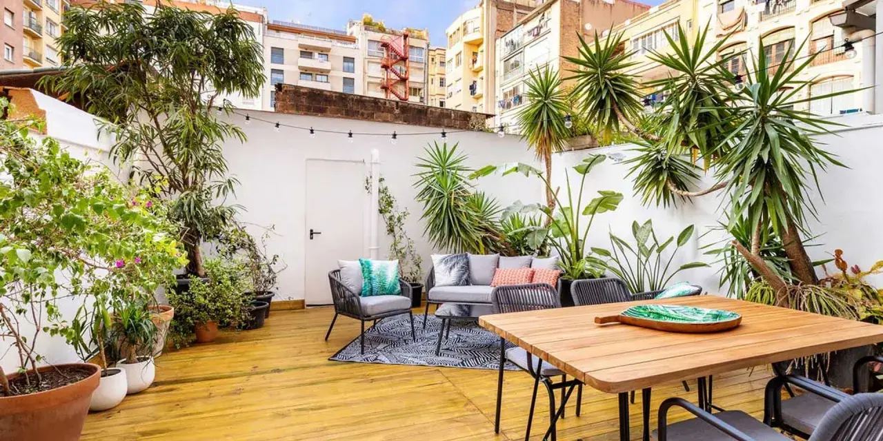 Un patio exterior pequeño transformado con una reforma: suelo de madera, muebles de exterior, plantas exuberantes y luces decorativas crean un oasis urbano.