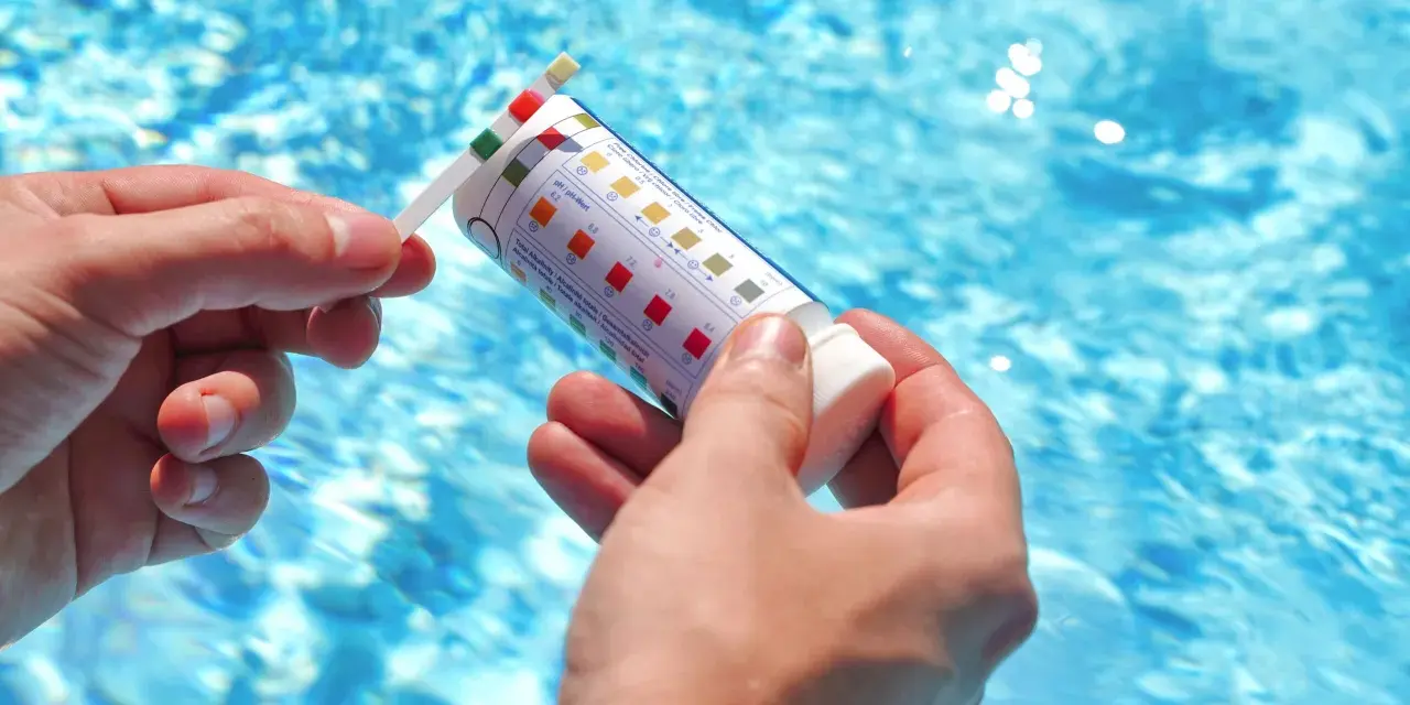 Manos sostienen una tira reactiva para medir el pH de la piscina. El agua azul y brillante de la piscina se ve al fondo.