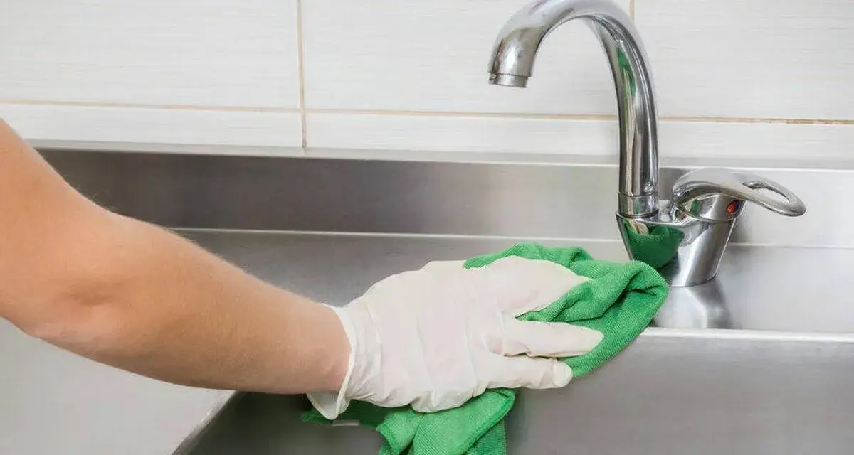 Mano con guante limpiando fregadero de acero inoxidable con paño verde.