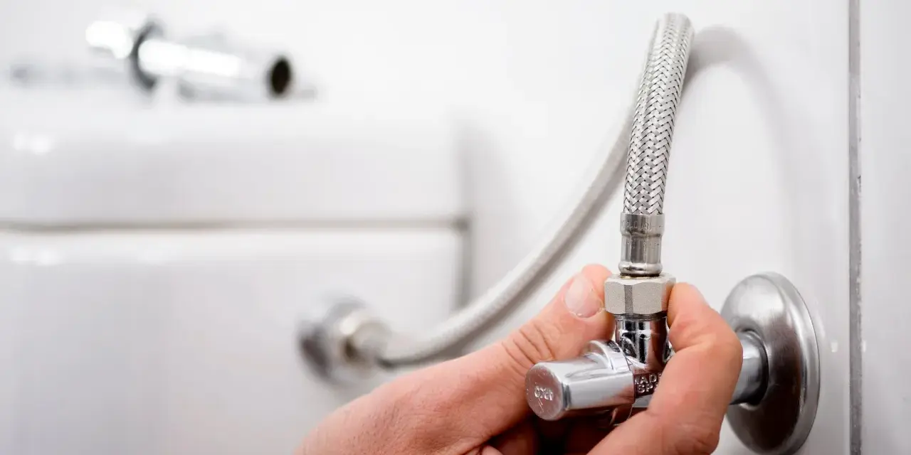 Mano ajustando uno de los tipos de llaves de paso de agua, conectada a una manguera flexible y al tanque de un inodoro.
