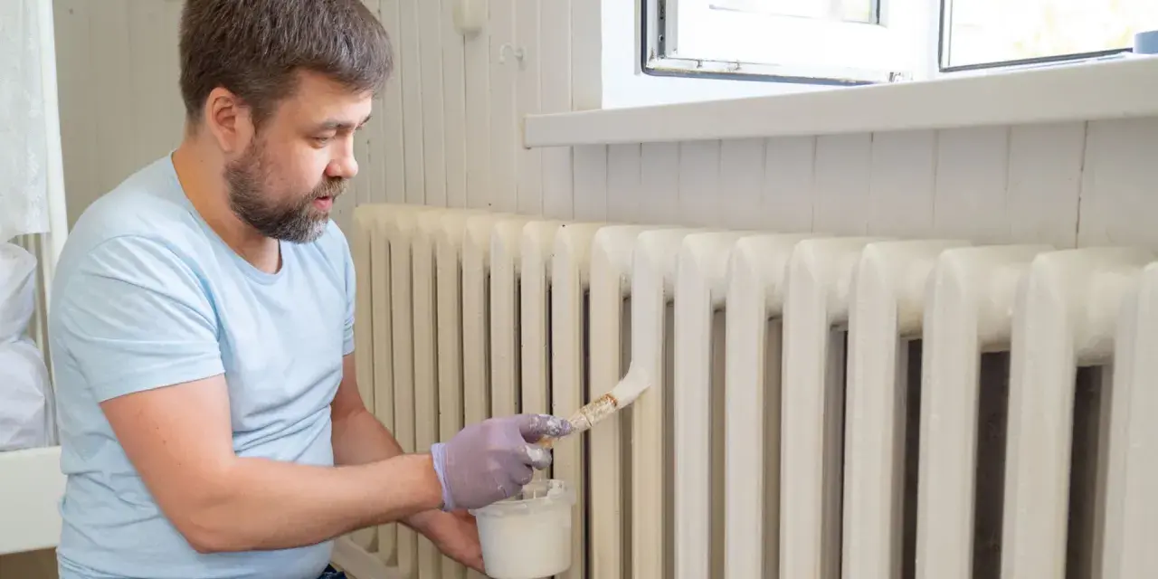 Hombre con barba pintando radiadores blancos con brocha y pintura.