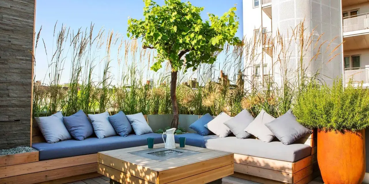Elegante decoracion terrazas aticos con sofás de madera, cojines grises y un centro de mesa con fuego. Hierbas altas y un árbol crean un oasis urbano.