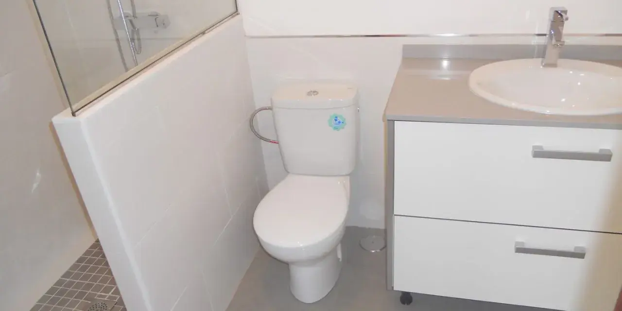 Baño moderno con ducha separada por muro de azulejos blancos y cristal. Inodoro y lavabo sobre mueble blanco.