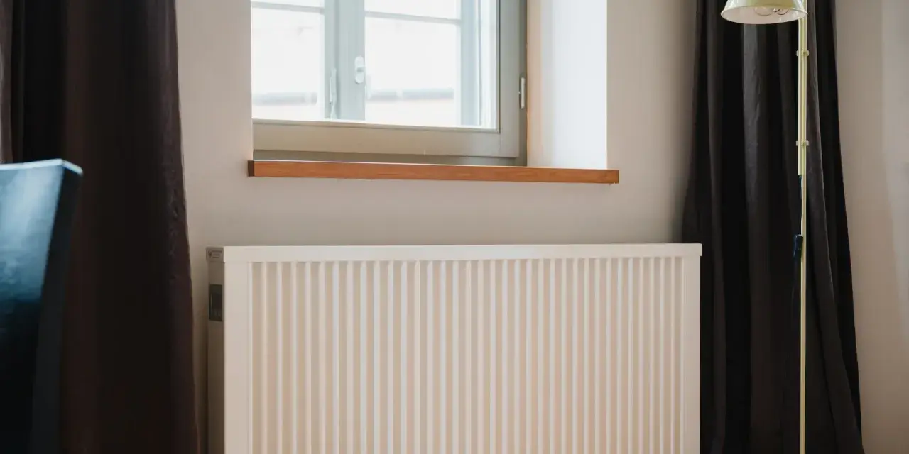 Radiador blanco bajo ventana, con cortinas oscuras y lámpara amarilla. El **calor azul** del invierno se combate con este sistema.