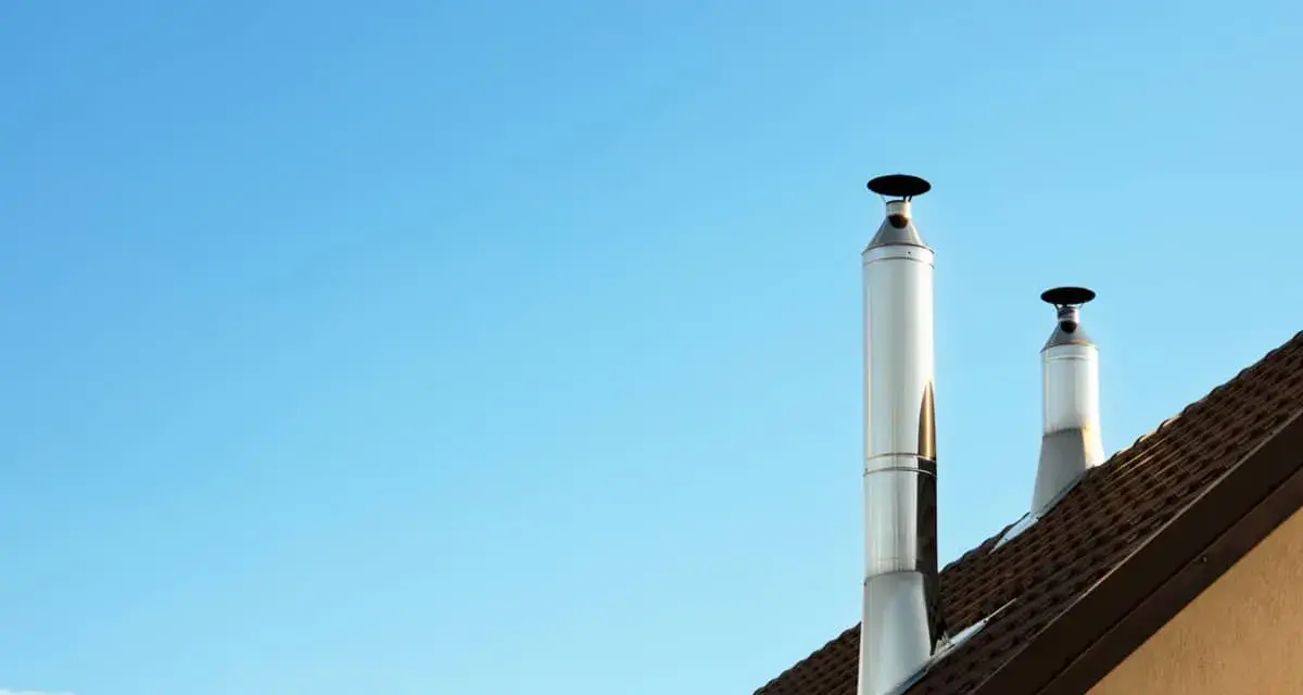 Dos tubos de chimenea metálicos se alzan sobre un tejado marrón contra un cielo azul claro. Es un día perfecto para aprovechar el calor del tubo de chimenea.