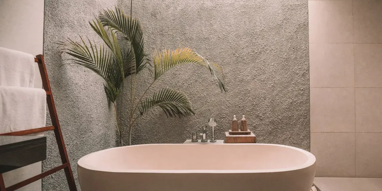 Un baño moderno con una bañera ovalada, toallas blancas en una escalera de madera y una planta de hojas verdes. La pared tiene textura y el suelo está cubierto de guijarros. ¡Listo para pintar la bañera y renovar el espacio!