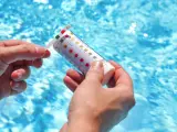 Manos sostienen una tira reactiva para medir el pH de la piscina. El agua azul y brillante de la piscina se ve al fondo.