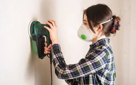 Mujer con mascarilla lijando paredes con una lijadora eléctrica.