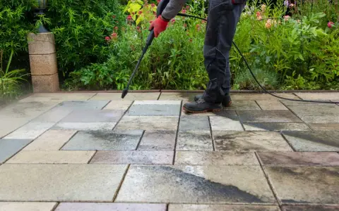 Persona con guantes rojos usa hidrolimpiadora para limpiar suelo de terraza exterior con baldosas de piedra.