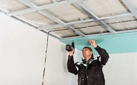Un obrero instala un **pladur techo** con una taladradora. La estructura metálica ya está fijada al techo original.