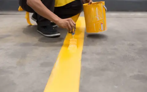 Persona pintando una línea amarilla sobre suelo de cemento sin pulir, con una lata de pintura al lado.